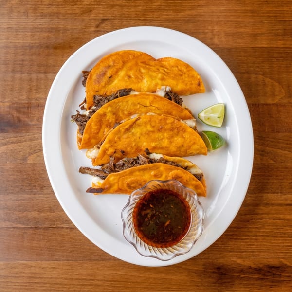 Four birria tacos with melted cheese and shredded beef, served with lime wedges and dipping sauce.