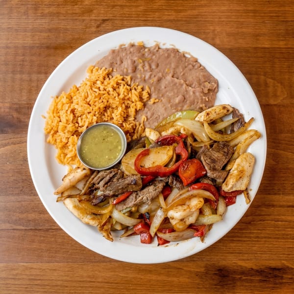 A white plate of fajitas with steak, chicken, shrimp, peppers, and onions, alongside rice, refried beans, and salsa.