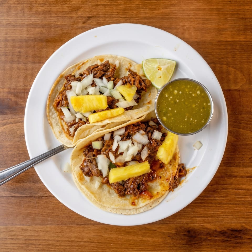 Two al pastor tacos with pineapple, onions, lime, and green salsa on a white plate.