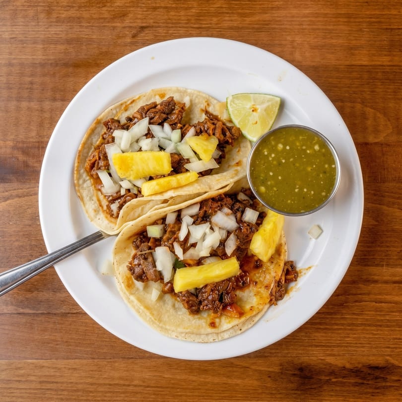 Two al pastor tacos with pineapple, onions, lime, and green salsa on a white plate.