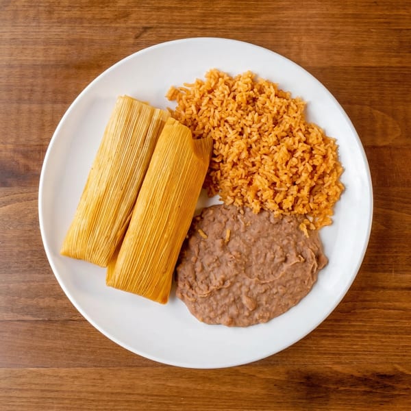 Two tamales, Spanish rice, and refried beans on a white plate on a wooden table.